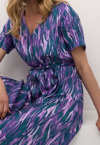 Robe multicolore avec une base turquoise ornée de motifs abstraits violets et lavande, manches courtes, col en V et un détail de taille nouée.