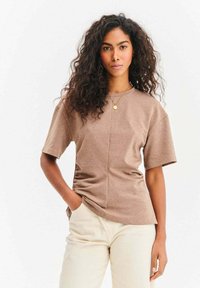 Kobieta o kręconych włosach, ubrana w beżową bluzkę z krótkim rękawem i kremowe spodnie, stoi z jedną ręką w kieszeni na jednolitym tle.