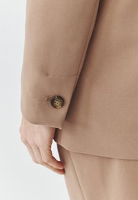 Dettaglio spalla del blazer beige, con un unico bottone marrone a quattro fori, vestibilità su misura, tessuto liscio e senza motivi visibili.