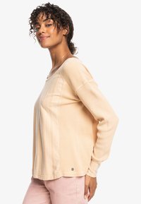Lichtbeige langemouwen ribbed top met een ontspannen pasvorm en een brede halslijn. Opmerkelijke details zijn onder andere subtiele verticale strepen en een klein logo nabij de zoom.