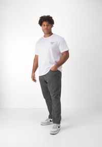 Bílý bavlněný tričko s logem "TOMMY JEANS", v kombinaci se šedými džínovými kalhotami a šedobílými teniskami Nike. Model stojí s rukama v kapsách.