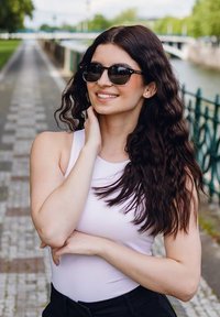 Černé sluneční brýle s kulatým rámem, které nosí žena s dlouhými, vlnitými tmavými vlasy, oblečená v bílém topu bez rukávů, stojící venku u cesty.