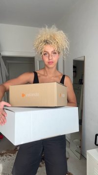 Două cutii ținute în mâini: una din carton maro cu logo-ul Zalando, cealaltă albă, rectangulară, robustă. Fundalul prezintă o oglindă și un decor ușor.