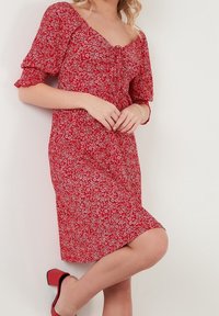 Robe fleurie rouge avec manches courtes bouffantes et encolure carrée. Présente un corsage ajusté et une taille cintrée, se terminant juste au-dessus du genou.