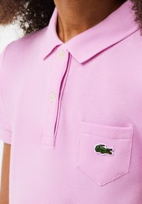 Růžová polo košile s texturovanou tkaninou, se dvěma knoflíky na límci a zeleným krokodýlím logem na levé kapse.