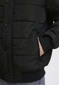 Czarna kurtka puchowa z przeszywanymi sekcjami, zatrzaskami, bocznymi kieszeniami i ściągaczami przy mankietach, wykonana z gładkiego, wodoodpornego materiału.