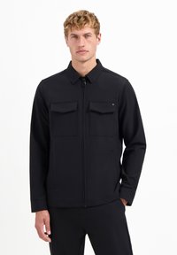 Jonge man in een zwarte zip-up jack met twee borstzakken, staand met één hand in zijn zak tegen een effen witte achtergrond.