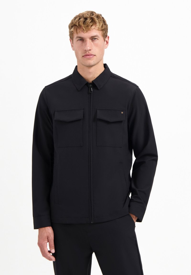 Jonge man in een zwarte zip-up jack met twee borstzakken, staand met één hand in zijn zak tegen een effen witte achtergrond.