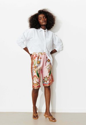 Femme aux cheveux bouclés portant une chemise blanche boutonnée, un short rose à motifs floraux paisley et des sandales à talons ouvertes couleur beige, debout contre un mur blanc.