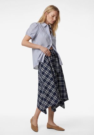 Blondynka poprawiająca niebieską kraciastą bluzkę z krótkim rękawem i granatową spódnicę midi w asymetryczną kratę, nosząca brązowe płaskie buty na białym tle.