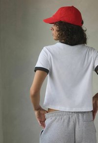 Bílý zkrácený t-shirt s černým lemem, doplněný šedými tepláky. Model má na hlavě červenou čepici, předvádí kudrnaté vlasy a minimalistické pozadí.