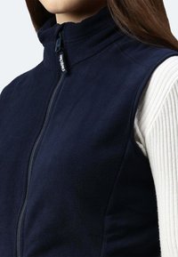 Marineblaue Fleece-Weste mit hohem Kragen, Reißverschluss und Logopatch. Verfügt über eine weiche Textur und eine figurbetonte Passform. Getragen über einem gerippten weißen Oberteil.