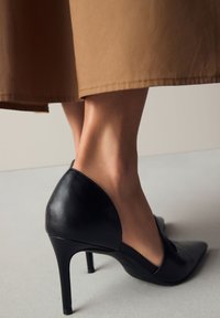 Czarny skórzany but na wysokim obcasie z zaostrzonym noskiem i wycięciami po bokach. Na neutralnym tle, w połączeniu z brązową spódnicą.