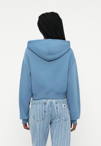 Casaco cropped azul feito de material macio com capuz, apresentando punhos e bainha canelados, combinado com jeans claros listrados de cintura alta.