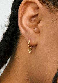 Goldene Creole mit einem kleinen Herzanhänger, mit glatter Oberfläche. Das Ohr des Modells ist teilweise sichtbar und zeigt die Textur und den Hautton.