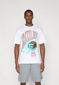 Λευκό βαμβακερό μπλουζάκι με γραφικά των Memphis Grizzlies σε τιρκουάζ και κόκκινο, κοντά μανίκια και λαιμόκοψη. Φορεμένο με γκρι σορτς.