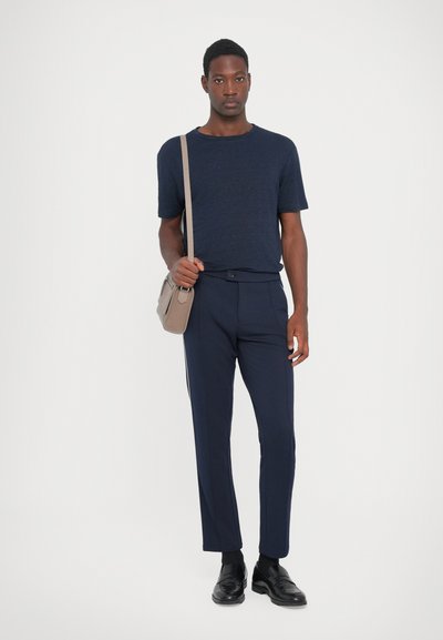 Marineblaues Kurzarm-T-Shirt kombiniert mit maßgeschneiderten marineblauen Hosen, die eine schmale Passform aufweisen, sowie einem passenden Gürtel. Eine beigefarbene Schultertasche rundet das Outfit ab.