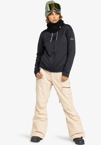 Black zip-up jacket with textured fabric, cream-colored snow pants with side zippers, black knitted neck warmer, green beanie, and ski goggles.