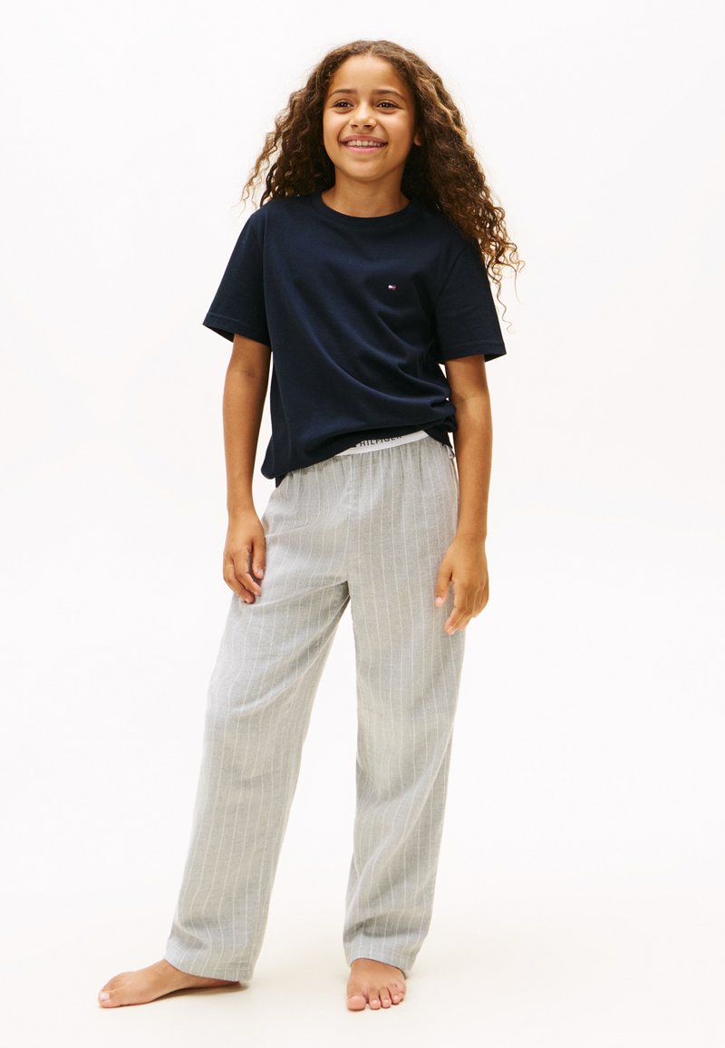 Bambino sorridente con i capelli lunghi e ricci, in piedi scalzo, con una maglietta nera e pantaloni grigio chiaro a righe, su uno sfondo bianco semplice.