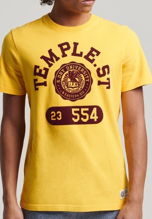 Żółta bawełniana koszulka z ciemnoczerwonym nadrukiem, zawierającym napis "TEMPLE.ST", godło orła oraz liczby "23 554". Krótkie rękawy, okrągły dekolt.