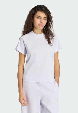 Ung kvinde med langt krøllet hår iført en lyse-lilla Adidas t-shirt og matchende bukser, står med den ene hånd i lommen.