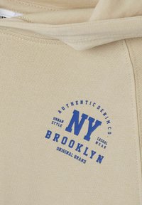 Sudadera beige con texto impreso en azul "NY BROOKLYN" y un diseño circular. Tejido suave con textura acanalada en los puños y el dobladillo.