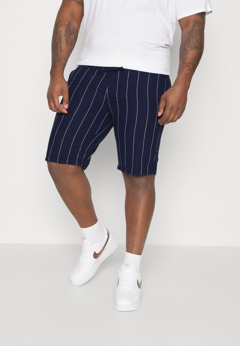 Homme portant un short bleu marine à rayures verticales blanches, un T-shirt blanc, des chaussettes blanches et des baskets blanches avec des logos métalliques en forme de swoosh.