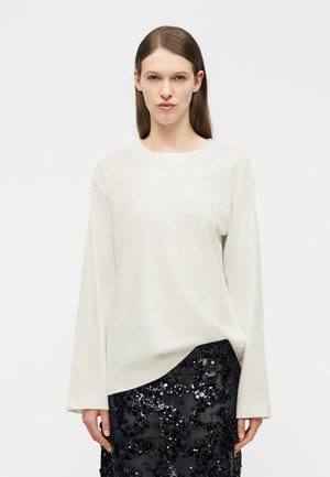 Žena s rovnými hnedými vlasmi má na sebe voľný biely top s dlhými rukávmi v kombinácii s čiernou sukňou s flitrami na jednoduchom svetlom pozadí.