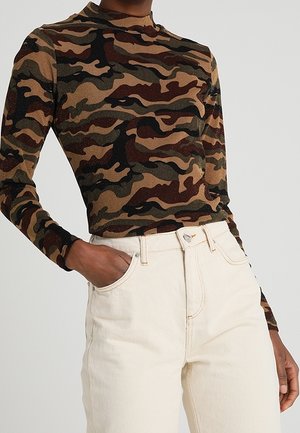 Personne portant un haut à manches longues camouflage marron et vert rentré dans un pantalon taille haute écru avec une main dans la poche.