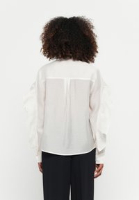 Hvit bluse med volangermuffe, lett stoff og detalj med bakfold. Avsluttet med knappemanchetter og buet kant.