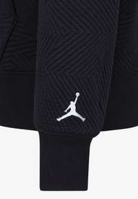 Černá textura mikiny s kapucí s herringbone vzorem, zdobená bílým logem Jumpman na rukávu a žebrovanými manžetami.