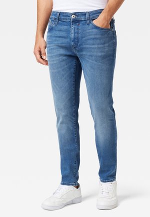 Man draagt slim-fit blauwe spijkerbroek en witte sneakers, staand met één hand in de zak tegen een effen achtergrond.