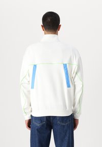 Balta zip-up striukė su mėlynais ir žaliais akcentais, laisvo kirpimo ir kontrastingais siūlais. Medžiaga atrodo lygi ir lengva.