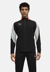 Giacca sportiva nera con accenti grigi e arancioni, chiusura frontale a zip completa, maniche lunghe e logo Puma sul lato sinistro del petto. Tessuto dalla consistenza liscia.