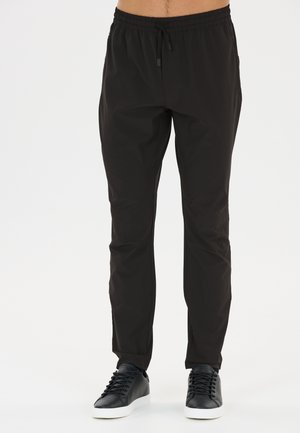 Homme portant un pantalon noir slim avec une taille élastique et des baskets noires à semelles blanches sur un fond uni.