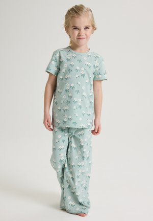 Jong meisje staand, gekleed in lichtgroene pyjama met wit en geel bloemenpatroon, tegen een effen lichte achtergrond.