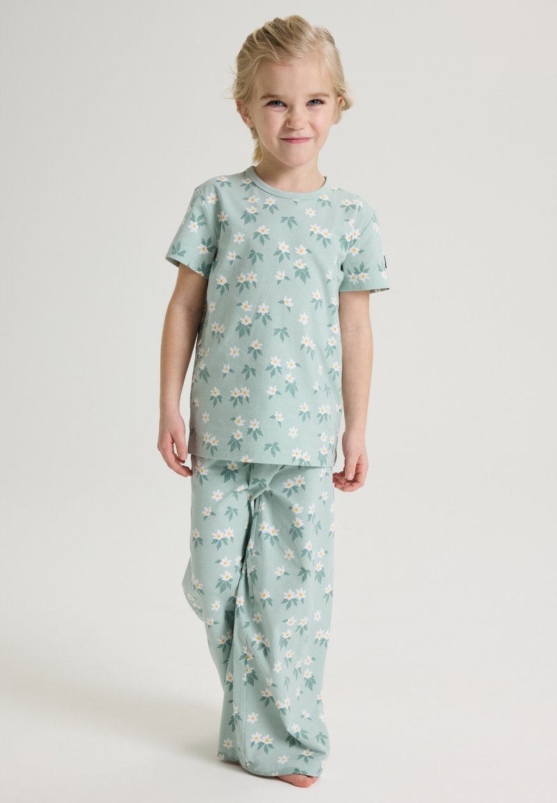 Young girl standing, wearing light green pajamas with white and yellow floral pattern, against plain light background.