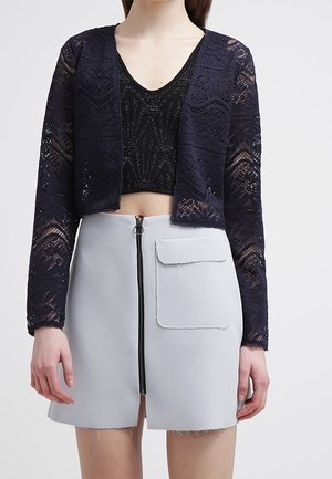 Kvinde iført en sort blonder crop cardigan, sort mønstret crop top og en lys grå mininederdel med lynlås og lomme foran.