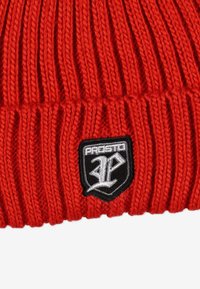 Czerwona dzianinowa czapka z prążkowaną fakturą i czarną naszywką z haftowanym logo przedstawiającym stylizowaną literę "P" na białym tle.