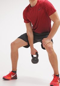 Camiseta deportiva de estampado rojo, pantalones cortos negros, sosteniendo una kettlebell. El calzado es rojo con detalles en negro. Enfoque en fitness y ambiente de entrenamiento.
