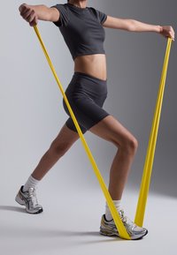 Haut de sport noir court et short. Bande de résistance jaune tendue entre les mains. Chaussures de sport argentées et jaunes avec chaussettes blanches.