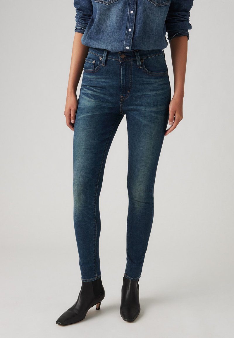 Skinny denim jeans in donkerblauw, met een hoge tailleband, subtiele vervagingdetails en een enkelhoogte snit, gecombineerd met zwarte haklaarzen.