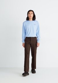 Camisa de manga larga azul claro con bordado en la parte frontal, combinada con pantalones marrones con bolsillos laterales y zapatos negros. Diseño simple y casual.