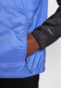 Giacca leggera blu con finitura testurizzata; presenta maniche nere, una tasca con zip e il marchio "WINDWALL" sul braccio.
