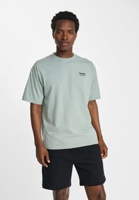 Ljusgrön bomullströja med korta ärmar, rund halsringning och en svart logotyp på vänster bröst, kombinerat med svarta shorts.