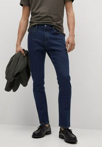 Vaqueros slim-fit de color azul oscuro hechos de denim, combinados con una camiseta oscura y una chaqueta verde, llevados con zapatos negros de cordones.