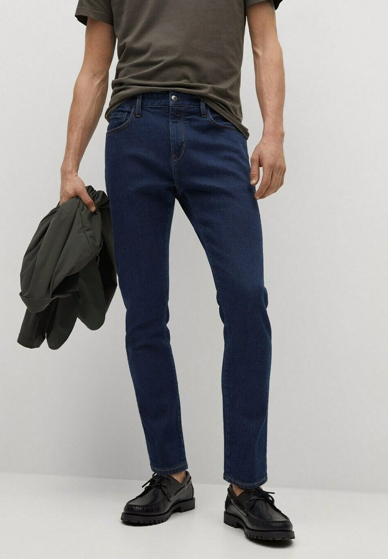 Vaqueros slim-fit de color azul oscuro hechos de denim, combinados con una camiseta oscura y una chaqueta verde, llevados con zapatos negros de cordones.
