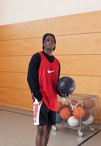 Rød Nike basketballtrøye over en svart langermet skjorte, svarte shorts, holder en basketball. Bakgrunnen har en vogn med ulike basketballer.