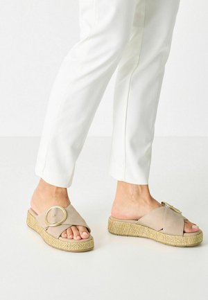 Fødder iført beige slidesandaler med stor guldbeslag og tekstureret platformsål, kombineret med hvide bukser.