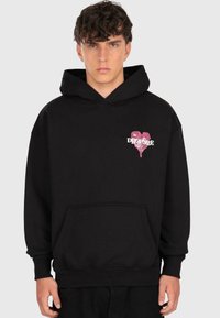 Young man with curly hair wearing a black hoodie with a pink dripping heart and the word "DROPSIZE" on the chest.